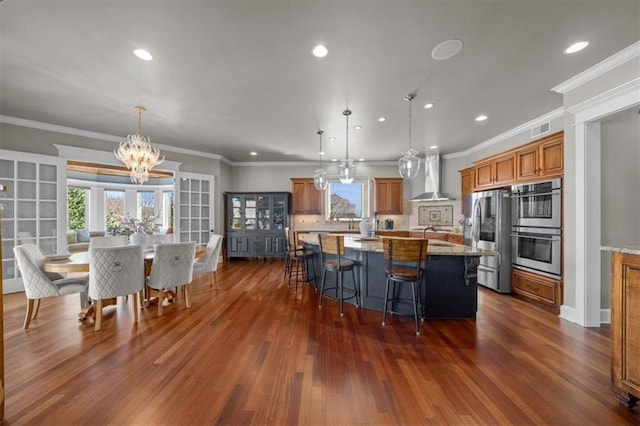 kitchen with a breakfast bar area, brown cabinetry, dark wood-style flooring, appliances with stainless steel finishes, and wall chimney range hood