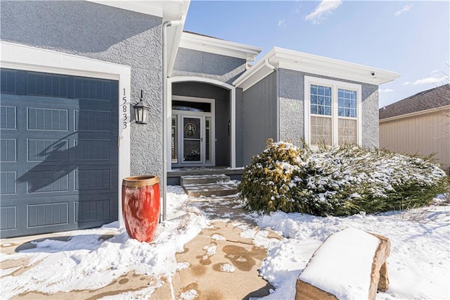 snow covered property entrance with an attached garage and stucco siding