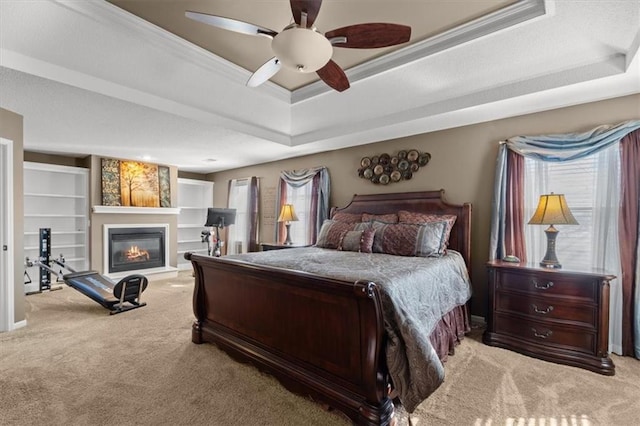 carpeted bedroom with ornamental molding, a glass covered fireplace, a raised ceiling, and ceiling fan