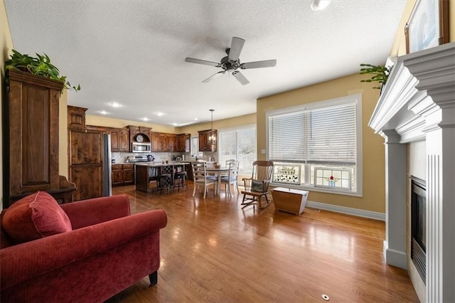 living room featuring a textured ceiling, a glass covered fireplace, baseboards, ceiling fan, and dark wood-style flooring