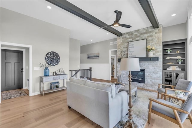 living room with ceiling fan, a large fireplace, built in shelves, beamed ceiling, and light wood-type flooring