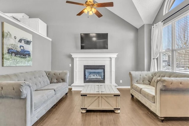 living room with a tiled fireplace, dark wood-type flooring, a ceiling fan, vaulted ceiling, and baseboards