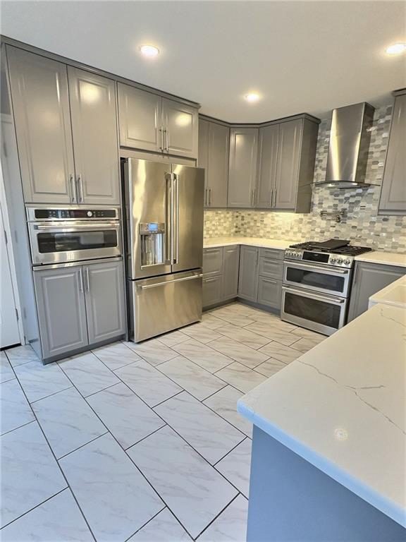 kitchen with gray cabinetry, wall chimney range hood, backsplash, and appliances with stainless steel finishes