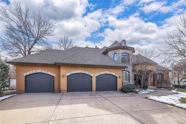 french country inspired facade with a garage, driveway, stone siding, roof with shingles, and stucco siding