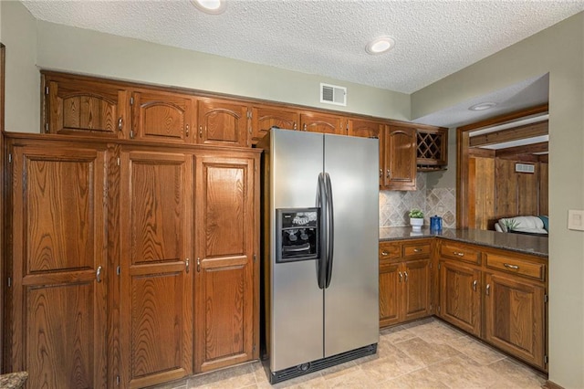 kitchen with brown cabinets, stainless steel refrigerator with ice dispenser, tasteful backsplash, dark countertops, and visible vents