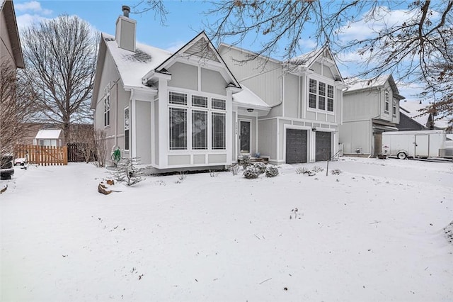 snow covered property with an attached garage, a chimney, and fence