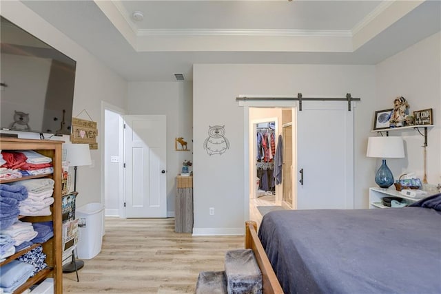 bedroom with a tray ceiling, a spacious closet, a barn door, light wood-style floors, and ornamental molding