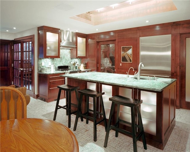 kitchen featuring a raised ceiling, an island with sink, light stone countertops, and wall chimney range hood