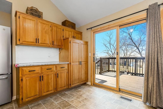 kitchen featuring brown cabinetry, visible vents, freestanding refrigerator, decorative backsplash, and vaulted ceiling