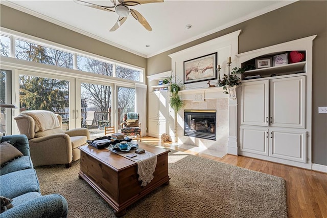 living area with baseboards, ceiling fan, a tiled fireplace, ornamental molding, and light wood-style floors