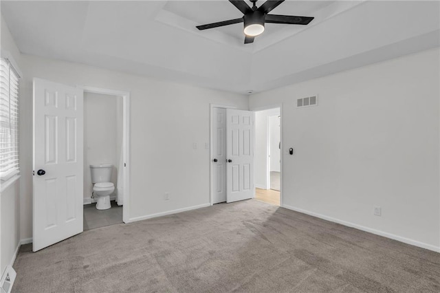 unfurnished bedroom with ceiling fan, ensuite bathroom, a raised ceiling, and light carpet