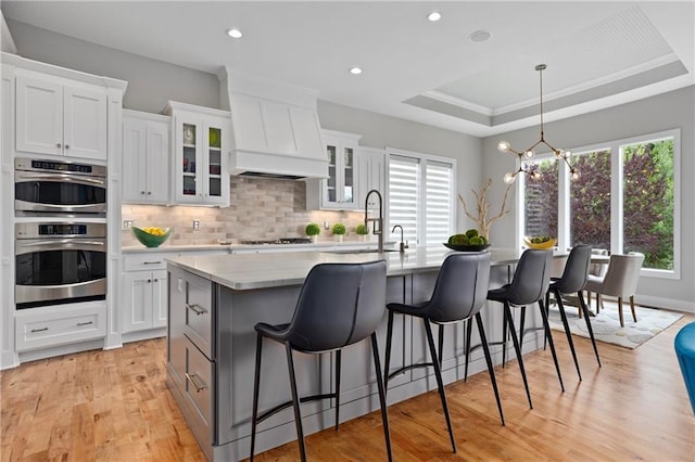 kitchen with backsplash, an island with sink, custom range hood, appliances with stainless steel finishes, and a raised ceiling