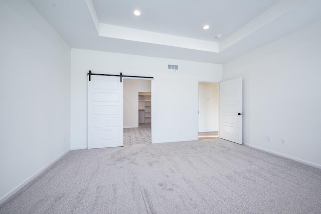 unfurnished bedroom with light carpet, a walk in closet, a raised ceiling, and a barn door