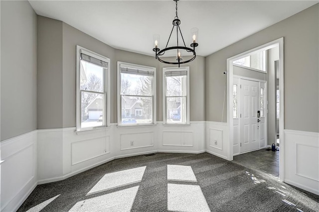 unfurnished dining area featuring wainscoting, dark carpet, a decorative wall, and an inviting chandelier