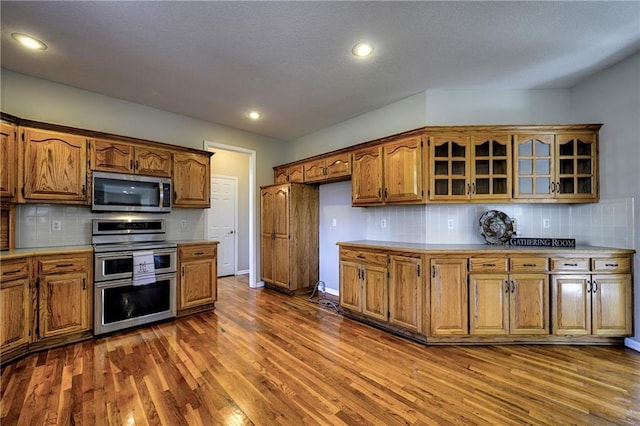 kitchen featuring stainless steel appliances, dark wood-style flooring, light countertops, brown cabinets, and glass insert cabinets