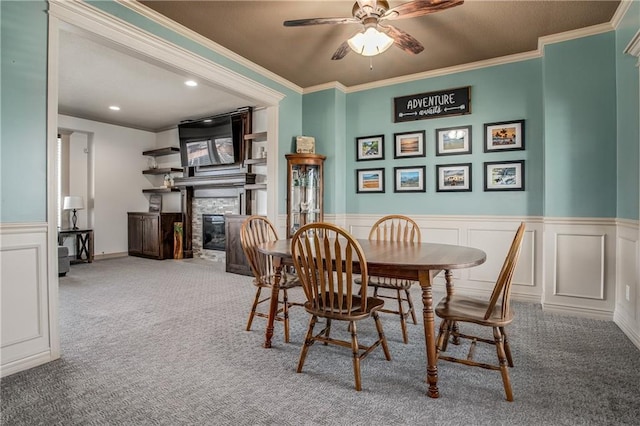 dining area featuring ornamental molding, carpet flooring, wainscoting, a stone fireplace, and a ceiling fan