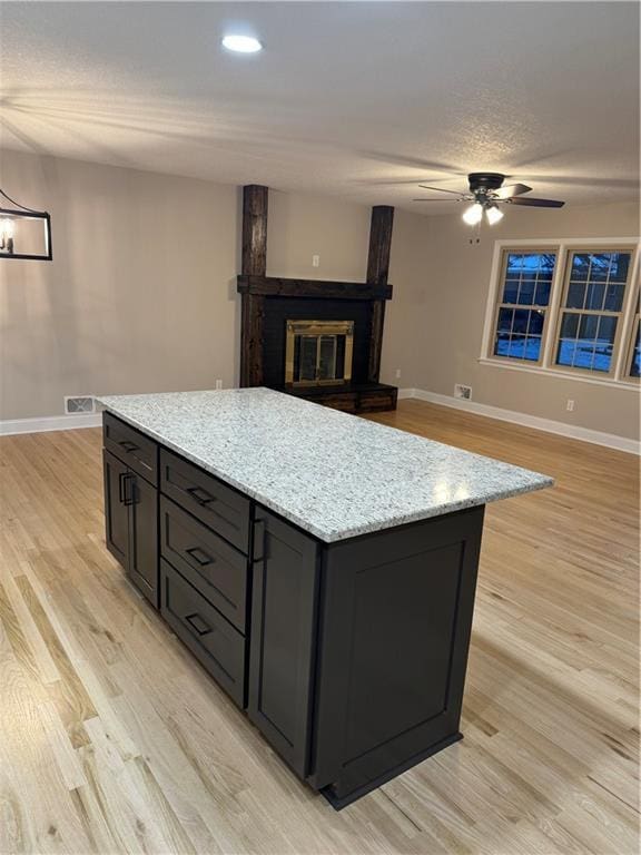 kitchen featuring light stone counters, a large fireplace, light hardwood / wood-style floors, ceiling fan with notable chandelier, and a kitchen island