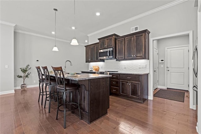 kitchen featuring a center island with sink, visible vents, stainless steel microwave, a kitchen breakfast bar, and dark wood-type flooring