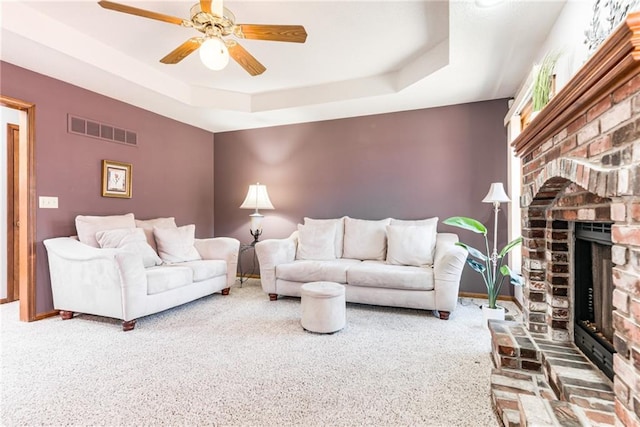 living area featuring visible vents, a ceiling fan, a tray ceiling, carpet floors, and a brick fireplace