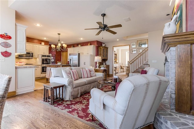 living area featuring stairway, light wood-style flooring, a fireplace, recessed lighting, and ceiling fan with notable chandelier