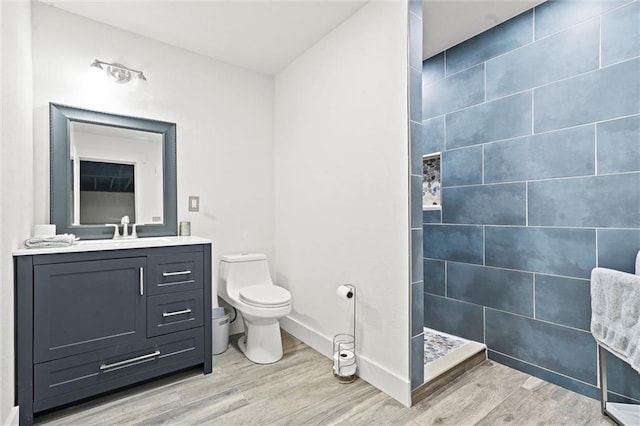 bathroom featuring toilet, a tile shower, vanity, wood finished floors, and baseboards