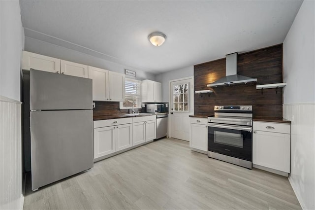 kitchen with light wood-style flooring, appliances with stainless steel finishes, white cabinets, a sink, and wall chimney exhaust hood