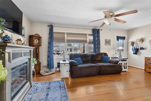 living room featuring a glass covered fireplace, a ceiling fan, baseboards, and wood finished floors