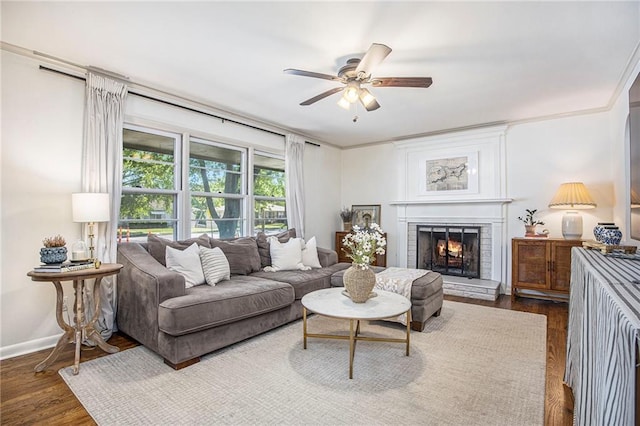 living room featuring a ceiling fan, a warm lit fireplace, dark wood-style flooring, baseboards, and crown molding