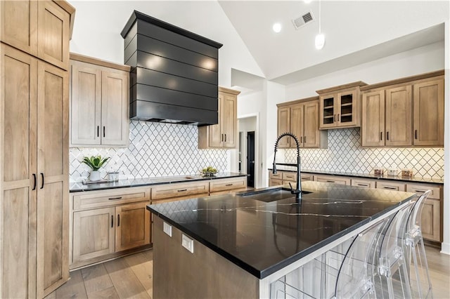 kitchen featuring vaulted ceiling, an island with sink, visible vents, wall chimney exhaust hood, and a sink