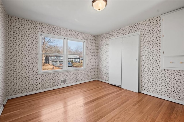 unfurnished bedroom featuring wallpapered walls, baseboards, visible vents, light wood-type flooring, and a closet