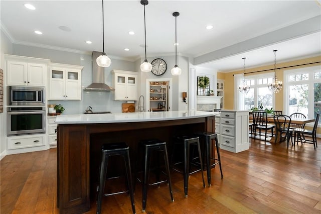 kitchen featuring crown molding, an island with sink, light countertops, wall chimney exhaust hood, and appliances with stainless steel finishes