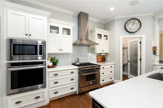 kitchen featuring wall chimney range hood, appliances with stainless steel finishes, light countertops, dark wood-type flooring, and ornamental molding