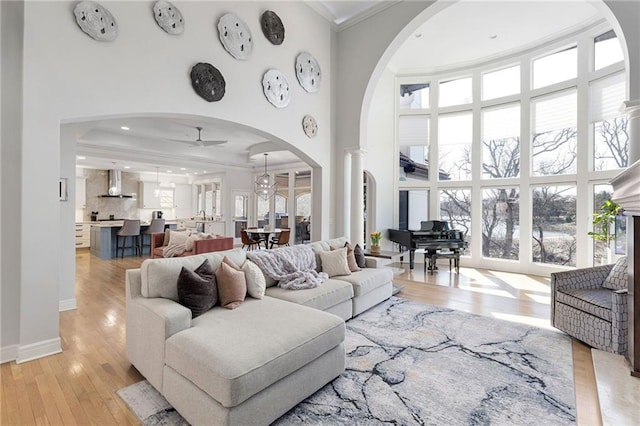 living area featuring ornamental molding, a ceiling fan, arched walkways, light wood-style floors, and a towering ceiling