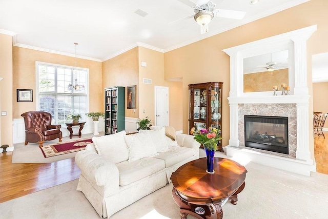 living area with ceiling fan with notable chandelier, crown molding, a high end fireplace, and light wood-style floors