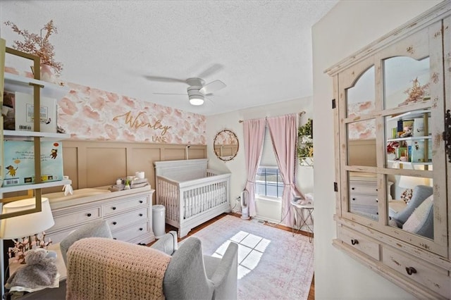 bedroom with a ceiling fan, wainscoting, a textured ceiling, a nursery area, and a decorative wall