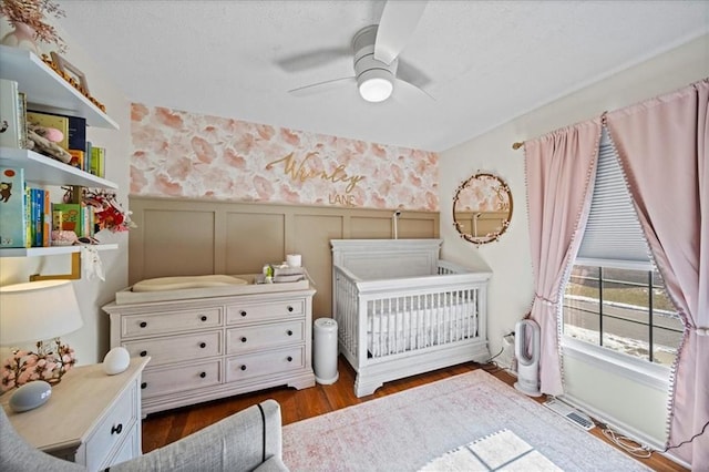 bedroom with a decorative wall, dark wood-type flooring, wainscoting, a crib, and wallpapered walls