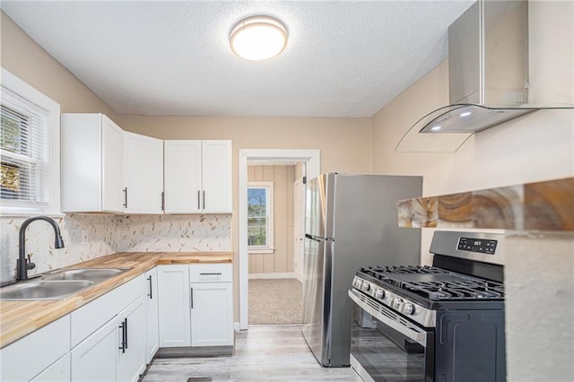 kitchen featuring sink, wall chimney range hood, stainless steel gas range oven, butcher block countertops, and white cabinets