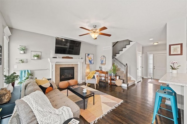 living area with visible vents, dark wood-type flooring, a glass covered fireplace, a ceiling fan, and stairs