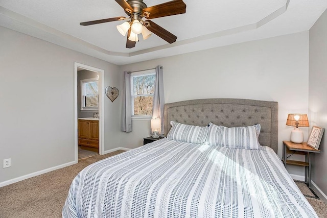 carpeted bedroom with ensuite bath, baseboards, a raised ceiling, and a ceiling fan