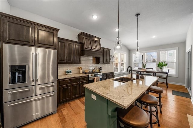 kitchen featuring stainless steel appliances, a kitchen island with sink, a sink, light stone countertops, and a kitchen bar