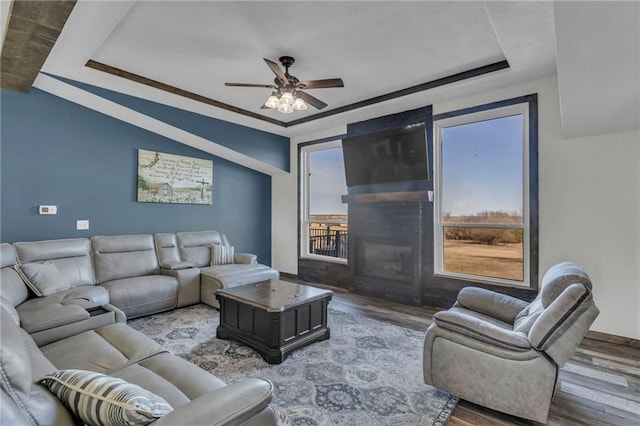 living room featuring ceiling fan, a tray ceiling, a glass covered fireplace, and wood finished floors