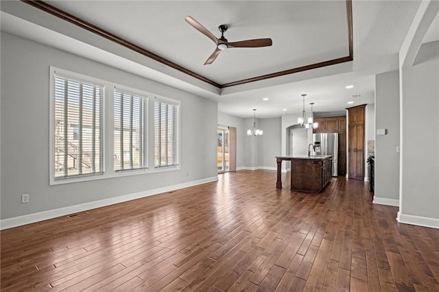 unfurnished living room with visible vents, ceiling fan with notable chandelier, a tray ceiling, dark wood-style floors, and baseboards