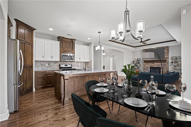 dining space with visible vents, ceiling fan with notable chandelier, a tray ceiling, a stone fireplace, and dark wood-style flooring