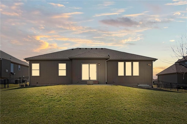 back of house at dusk with a lawn, fence, and a shingled roof