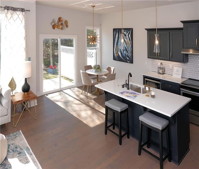 kitchen featuring a kitchen island with sink, under cabinet range hood, light countertops, hanging light fixtures, and stainless steel electric range oven