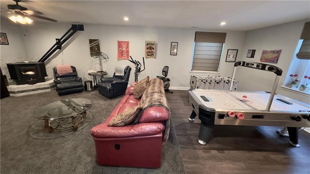 rec room featuring ceiling fan, recessed lighting, dark wood-type flooring, baseboards, and a wood stove