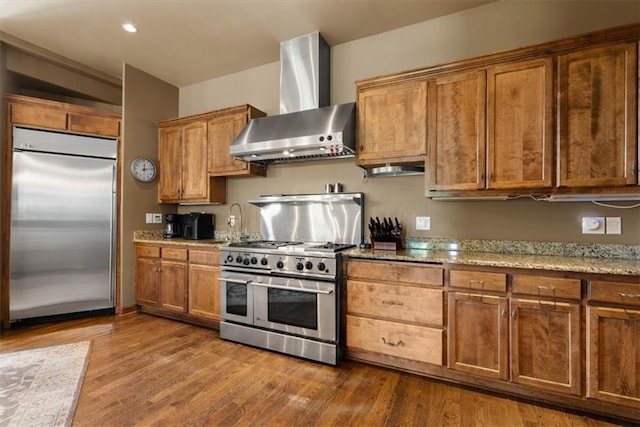 kitchen featuring wall chimney range hood, high end appliances, wood finished floors, and brown cabinetry