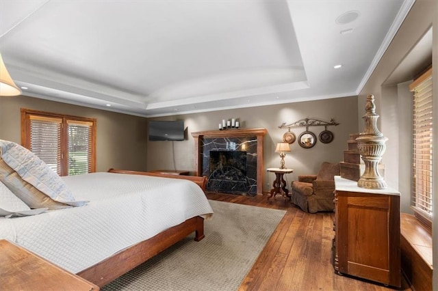 bedroom with recessed lighting, a premium fireplace, wood finished floors, a raised ceiling, and crown molding