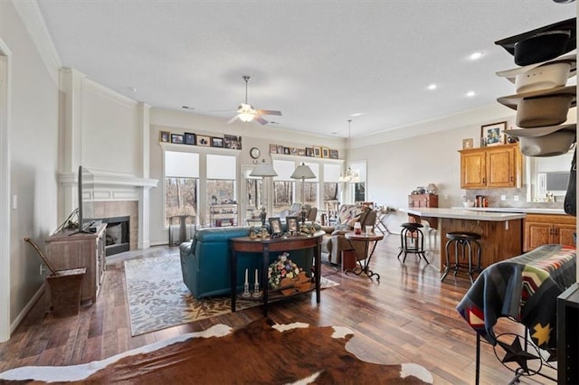 living area featuring a tile fireplace, dark wood-style flooring, a ceiling fan, baseboards, and crown molding