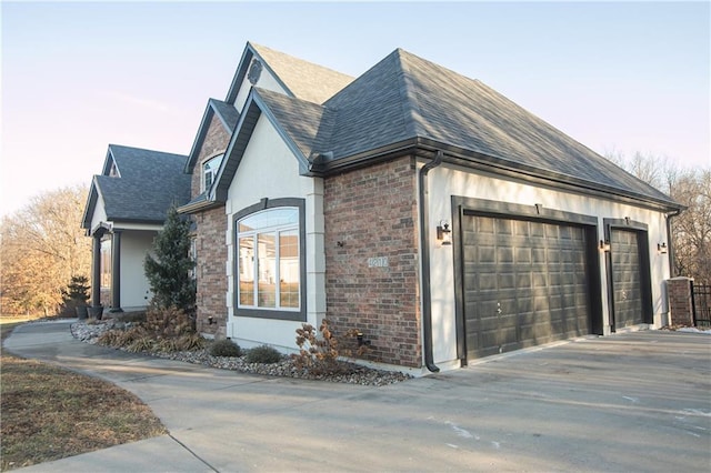 view of property exterior featuring an attached garage, brick siding, a shingled roof, driveway, and stucco siding